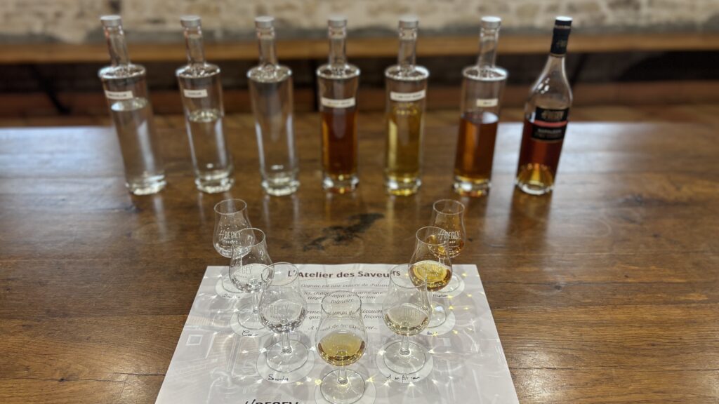 Row of six tall bottles with clear to amber liquids lined up on a wooden bar, with tasting glasses in the foreground and a printed sheet beneath.