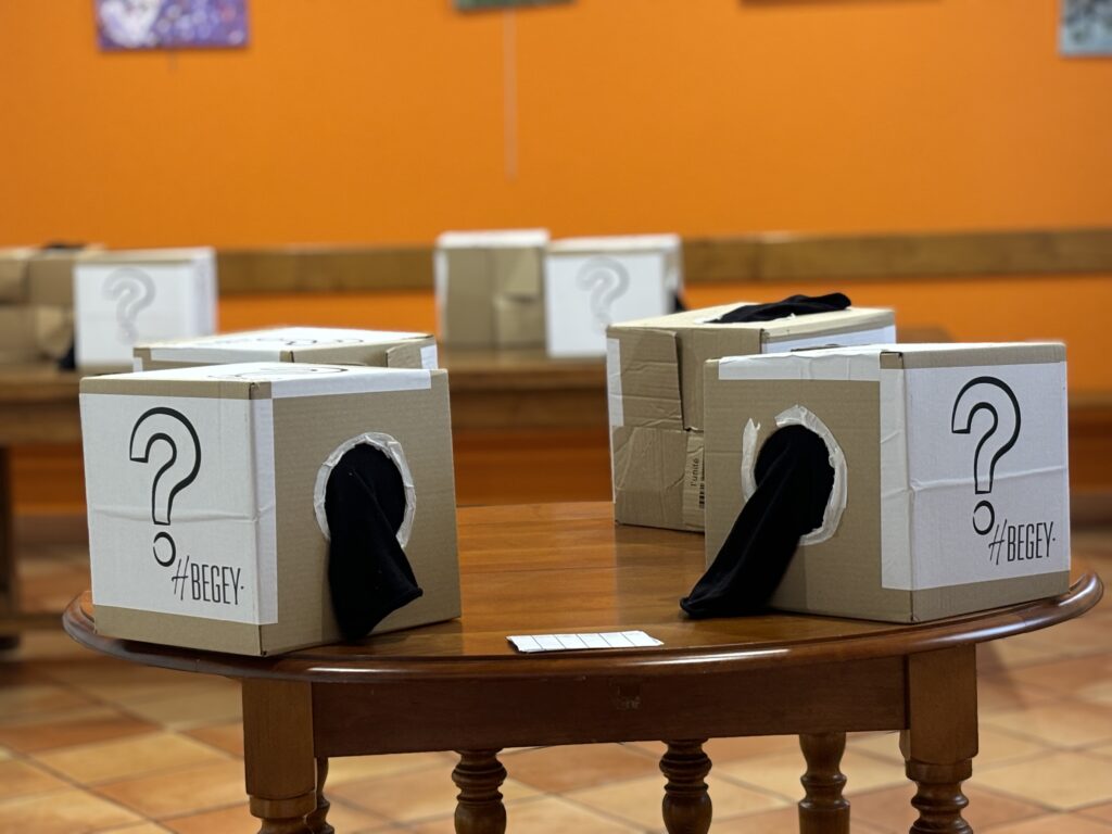 Cardboard boxes with large question marks placed on a round wooden table in a room with an orange wall, suggesting mystery or guessing activity.